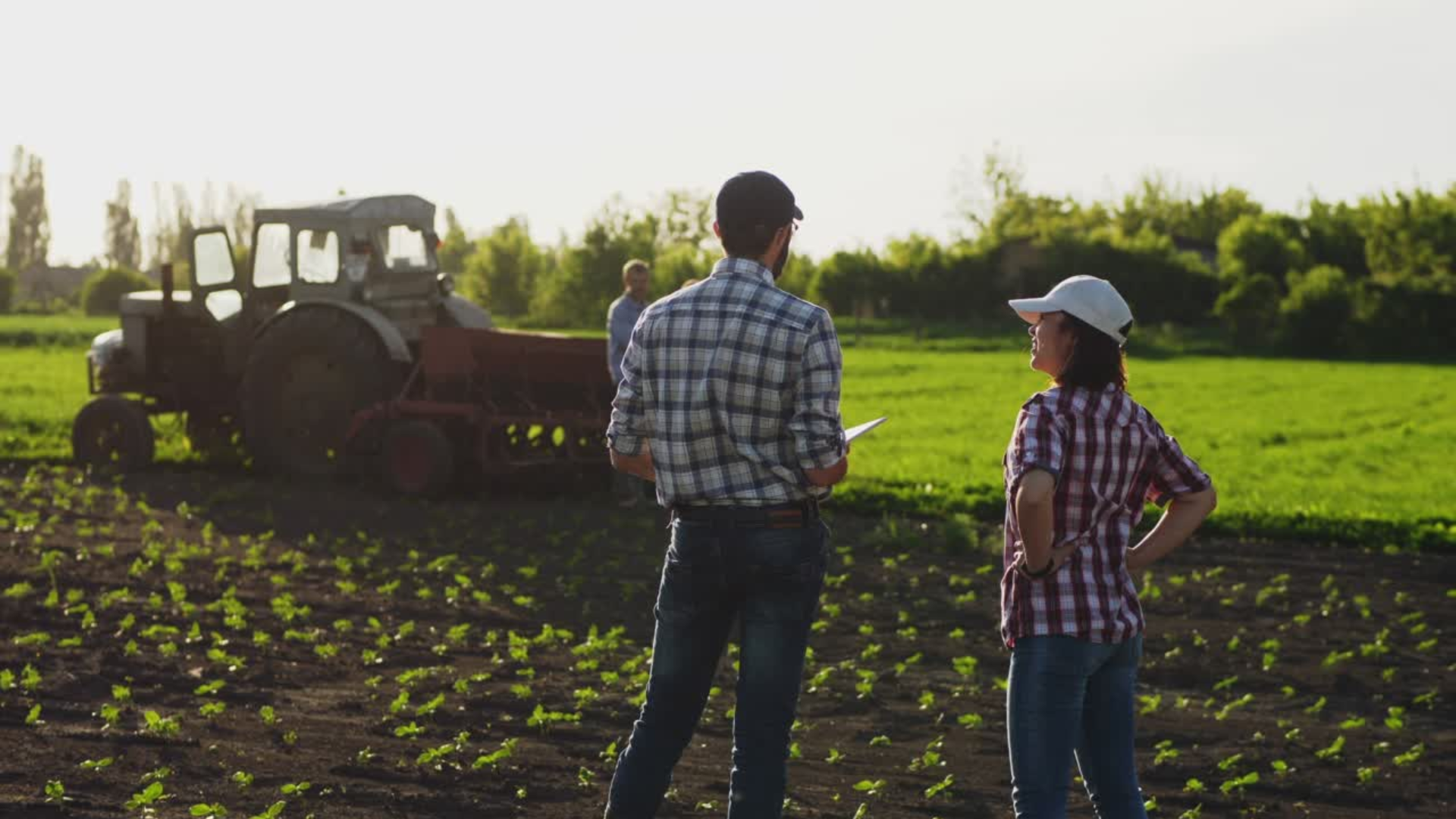 A farmer and a conservation planner talk in a field, a tractor driving in the background.