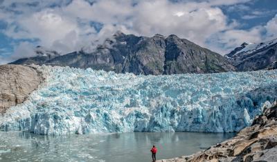 A person stands in front the glacier-blue LeConte Glacier. Mountains rise high in the background. 