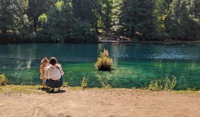 : A mother and young daughter face away from the camera, looking into a blue-green lake. The mother crouches next to her daughter. Across the lake are tall evergreen trees.
