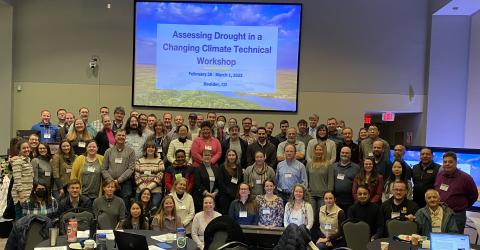 A group of people standing in front of a projector screen that reads "Assessing drought in a changing climate technical workshop - February 28-March 1, 2023 - Boulder, CO"