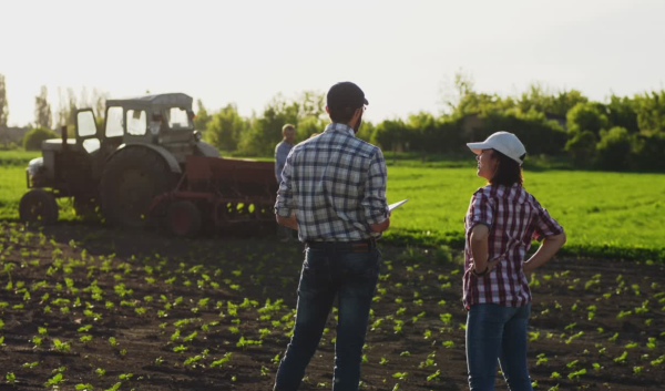 A farmer and a conservation planner talk in a field, a tractor driving in the background.